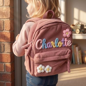May include: A dusty rose corduroy backpack featuring the name Charlotte in colorful, raised yarn lettering. The bag includes a top handle, a front zippered pocket, and decorative fabric flower appliqués in pink and white with yellow centers.