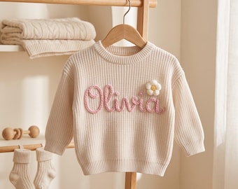 May include: A cream-colored long-sleeve knit sweater featuring the name Olivia embroidered in pink yarn across the chest, accented by a small white and yellow three-dimensional flower applique on the upper right side.