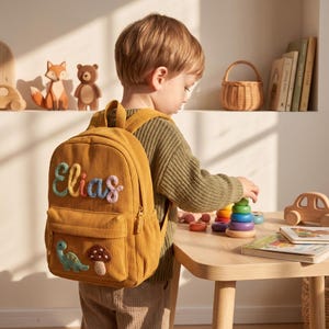 Puede incluir: Mochila de pana amarillo mostaza con el nombre "Elias" bordado en hilo colorido. Cuenta con un aplique de dinosaurio y seta. La mochila tiene un bolsillo frontal y cierre de cremallera. Un niño lleva la mochila.