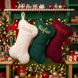 May include: Three bone-shaped Christmas stockings made of cable-knit fabric in cream, dark green, and burgundy. Each stocking features a hanging loop and is embroidered with a name in gold thread: Mochi on the cream, Rufus on the green, and Poppy on the burgundy.