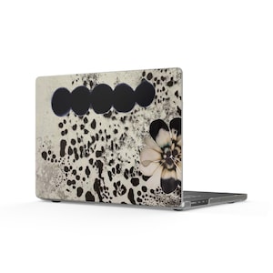May include: A laptop featuring a protective skin with an off-white background, a pattern of scattered black ink-like splatters, a horizontal row of five solid black circles, and a large stylized flower graphic with black and white petals.