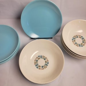 Op de afbeelding: Een verzameling keramisch servies, waaronder lichtblauwe borden en crèmekleurige kommen. De kommen hebben een decoratief ringpatroon in blauw, bruin en groen. De borden zijn gestapeld en op een wit oppervlak geplaatst.