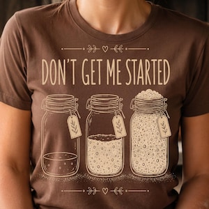 May include: A brown t-shirt featuring the text "DON'T GET ME STARTED" above a graphic of three glass jars with tags, showing varying levels of sourdough starter. The design includes small heart and leaf accents, printed in a light cream color.