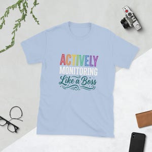 Könnte beinhalten: Hellblaues T-Shirt mit dem Text "ACTIVELY MONITORING Like a Boss" in bunten Buchstaben. Das Shirt hat kurze Ärmel und einen Rundhalsausschnitt. Eine Kamera, eine Brille, eine Geldbörse und ein Telefon sind im Hintergrund zu sehen.
