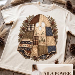 Może przedstawiać: Kremowy t-shirt z centralną grafiką ducha wykonanego z różnych wzorów tkanin patchworkowych, w tym w kratę, splotów warkoczowych i kwiatowych nadruków, otoczonego obwódką z liści dębu i szyszek. Tekst głosi AILA POWER.