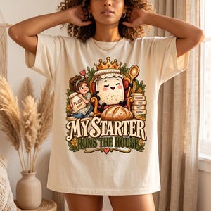 May include: A cream-colored t-shirt featuring a graphic of a sourdough starter jar wearing a crown, sitting in a chair with a wooden spoon. A person holds a flour bag nearby. Text reads: MY STARTER RUNS THE HOUSE, FEEDING, RISE, REST, REPEAT.