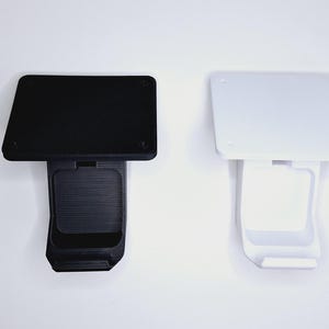 May include: Two 3D-printed phone holders, one black and one white, are shown against a white backdrop. Each holder has a rectangular top and a textured, curved base with a slot for a phone.