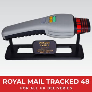 May include: A grey and black handheld phaser prop mounted on a black display stand. The phaser features yellow and green buttons, a red and black emitter tip, and a plaque reading PHASER TYPE II DUSTBUSTER YEARS OF SERVICE 2364-2370.