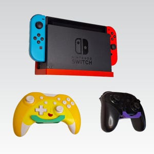 May include: A black Nintendo Switch console with blue and red Joy-Con controllers, and a red dock. Two game controllers are also shown: a yellow and white controller, and a black controller with a purple accent.