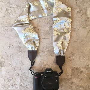 Pode incluir: Uma correia de câmera feita de tecido cinza claro com um padrão floral em amarelo e azul. A correia tem pontas de couro marrom e está presa a uma câmera preta com uma lente preta. A câmera tem a palavra "Nikon" impressa nela.
