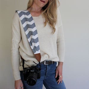 May include: A camera strap featuring a grey and white zigzag pattern. The strap is made of fabric with brown leather ends. A black camera is attached to the strap. The strap is worn over the shoulder.