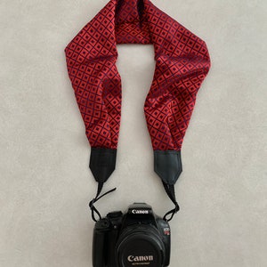 Pode incluir: Uma alça de câmera com padrão vermelho e preto com uma câmera Canon EOS Rebel T1i preta anexada.