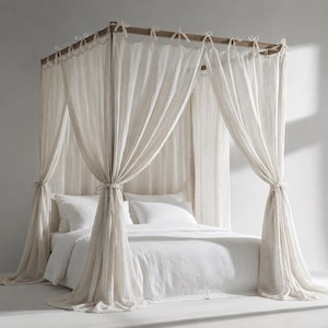 May include: A four-poster bed frame constructed from dark wood, featuring sheer, off-white fabric curtains draped from the top rails and tied at the posts. The bed is dressed with white pillows and a white duvet, creating a layered, soft appearance.