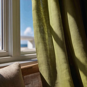 May include: A semi-sheer olive green curtain panel with a visible woven texture hangs vertically. The fabric is draped in soft folds, allowing light to pass through the weave, creating highlights and shadows across the surface of the material.