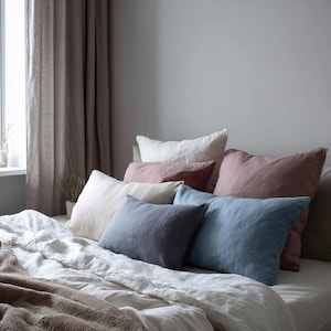 May include: A collection of rectangular and square throw pillows in various colors including dusty rose, slate blue, charcoal gray, and off-white, arranged on a bed with a white linen duvet and a textured brown throw blanket at the foot of the bed.