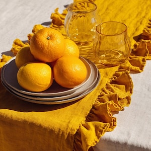 May include: A still life composition with a stack of grey plates holding oranges. Two clear, textured glass tumblers and a yellow linen table runner with ruffled edges are also present. The oranges are a vibrant orange colour.