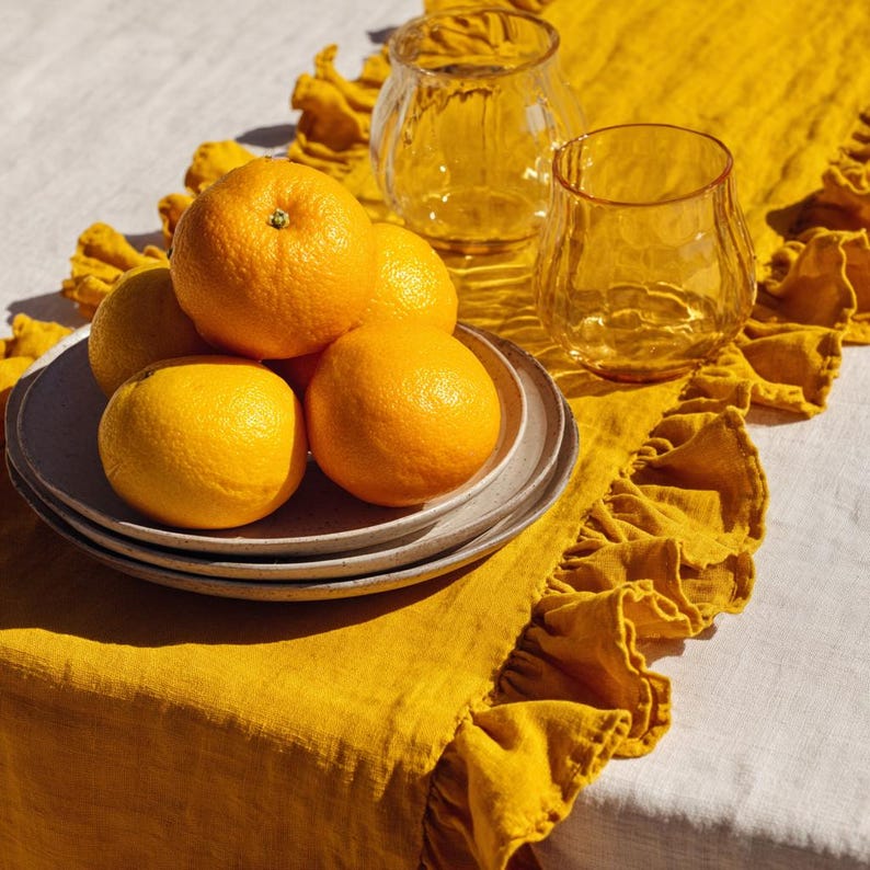 May include: A still life composition with a stack of grey plates holding oranges. Two clear, textured glass tumblers and a yellow linen table runner with ruffled edges are also present. The oranges are a vibrant orange colour.