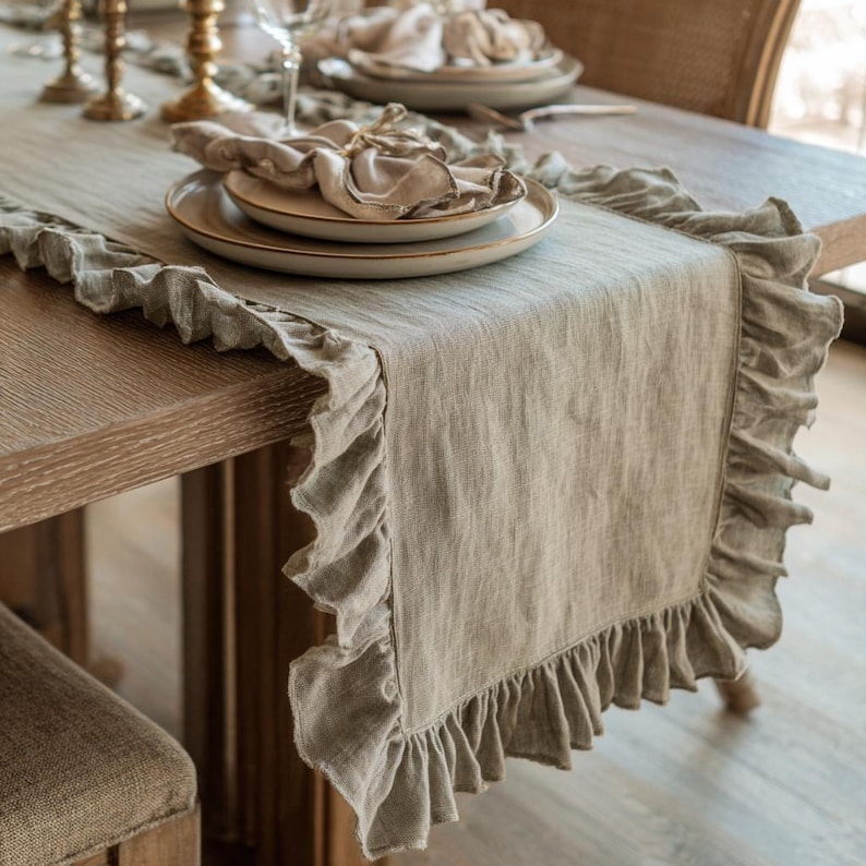 May include: A neutral linen table runner with a ruffled trim. The runner is laid on a wooden table, set with plates, napkins, and cutlery. The table setting has a rustic, elegant aesthetic, suitable for a dining room or special occasion.