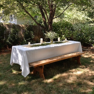 May include: A long wooden bench with a matching table covered in a white tablecloth, set outdoors under a tree. The table is decorated with candles and greenery, creating a rustic and inviting setting for an outdoor event.