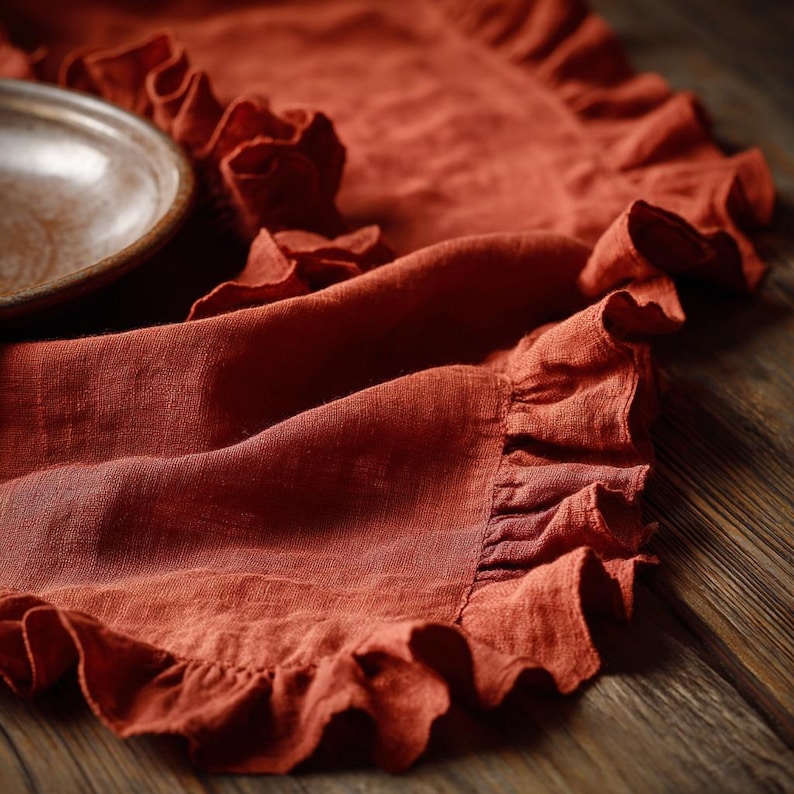 May include: A terracotta-coloured linen table runner with a ruffled edge. A section of a wooden plate is visible on the left. The runner is draped over a wooden surface, creating a rustic and textured aesthetic.