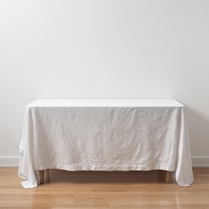 May include: A rectangular table covered with a solid white linen tablecloth that drapes over the sides. The fabric has a visible woven texture and soft folds, extending down to the wooden legs of the table, creating a clean and simple appearance.