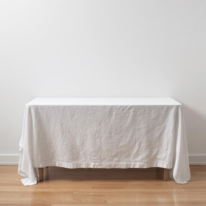 May include: A rectangular table covered with a solid white linen tablecloth that drapes over the sides. The fabric has a visible woven texture and soft folds, extending down to the wooden legs of the table, creating a clean and simple appearance.