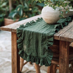 May include: A dark green linen table runner with frilled edges on a rustic wooden table. A beige vase with greenery and a stack of plates are on the table. The table is outdoors.