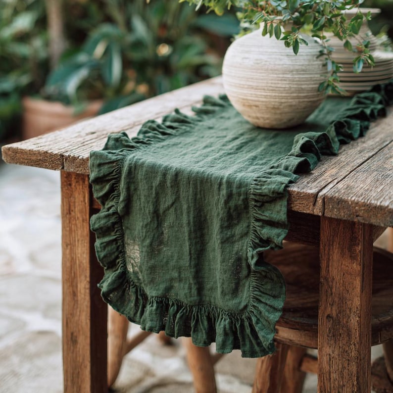 May include: A dark green linen table runner with frilled edges on a rustic wooden table. A beige vase with greenery and a stack of plates are on the table. The table is outdoors.