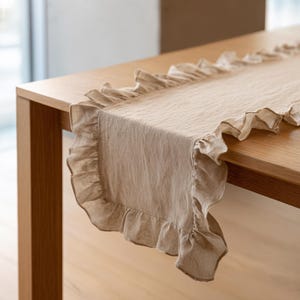 May include: A beige linen table runner with frilled edges. The runner is laid on a wooden table, highlighting its textured fabric and decorative trim. This item is suitable for home decor and special events.
