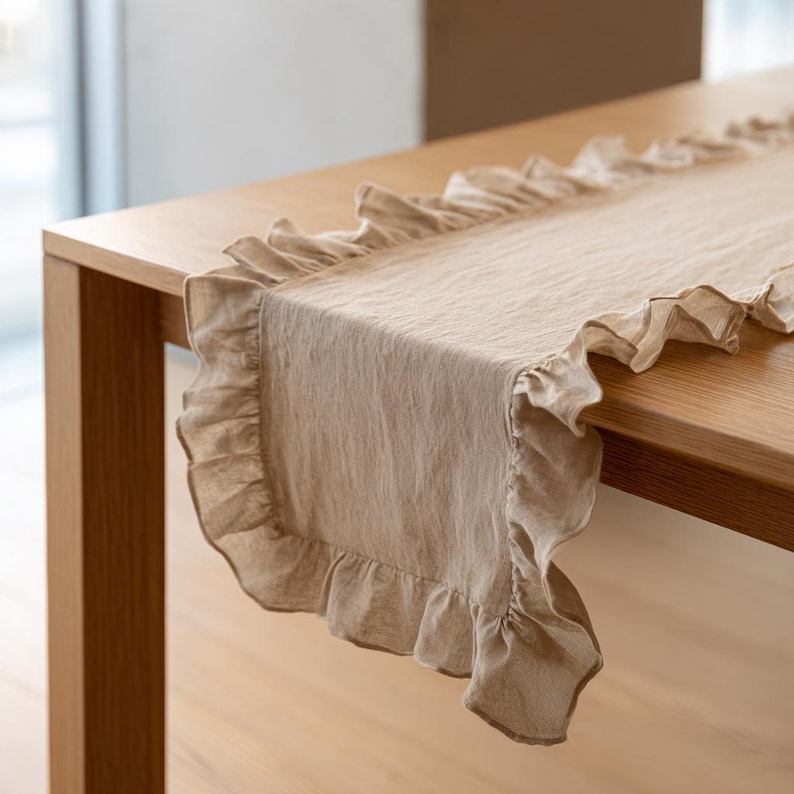 May include: A beige linen table runner with frilled edges. The runner is laid on a wooden table, highlighting its textured fabric and decorative trim. This item is suitable for home decor and special events.