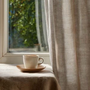 May include: A beige ceramic coffee cup with a matching saucer sits on a linen-covered surface near a window. The cup has a simple design with a slightly darker rim. A beige curtain hangs to the right, and a blurred view of greenery is visible through the window.