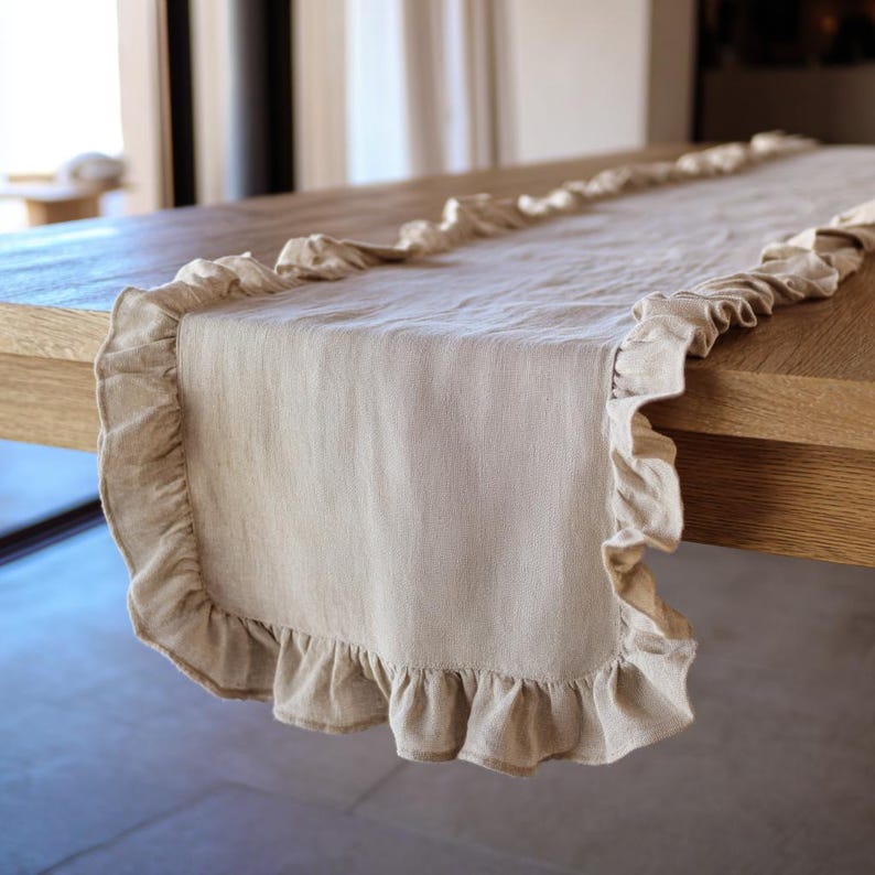 May include: A beige linen table runner with a ruffled edge. The runner is laid on a wooden table, highlighting its textured fabric and decorative trim. This elegant piece adds sophistication to any dining setting.
