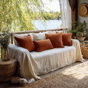 May include: A white linen sofa with four rust-colored throw pillows sits on a wooden deck. The sofa is covered with a white linen throw. A woven rug and a glass vase with dried flowers are in the foreground. The background shows a lake and trees.