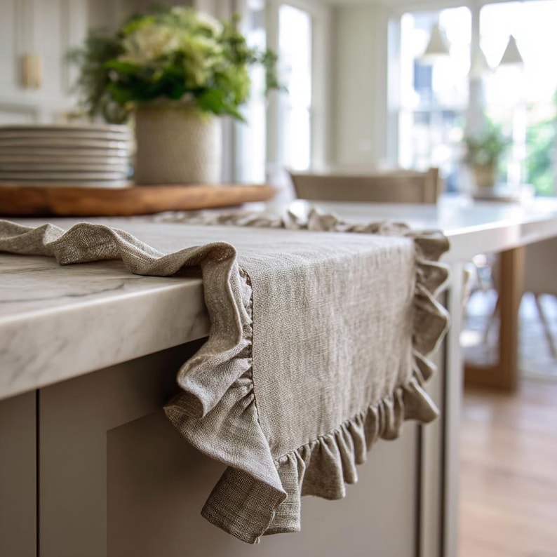 May include: A beige linen table runner with a ruffled trim. The runner is displayed on a kitchen countertop, highlighting its texture and design. The background features a stack of plates and a vase of flowers.
