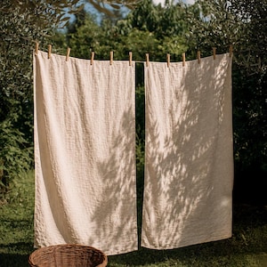 May include: Two rectangular, off-white linen towels hang side-by-side from a horizontal clothesline secured with wooden clothespins. A round, woven brown wicker basket sits on the ground below the hanging fabric, which features a visible weave.
