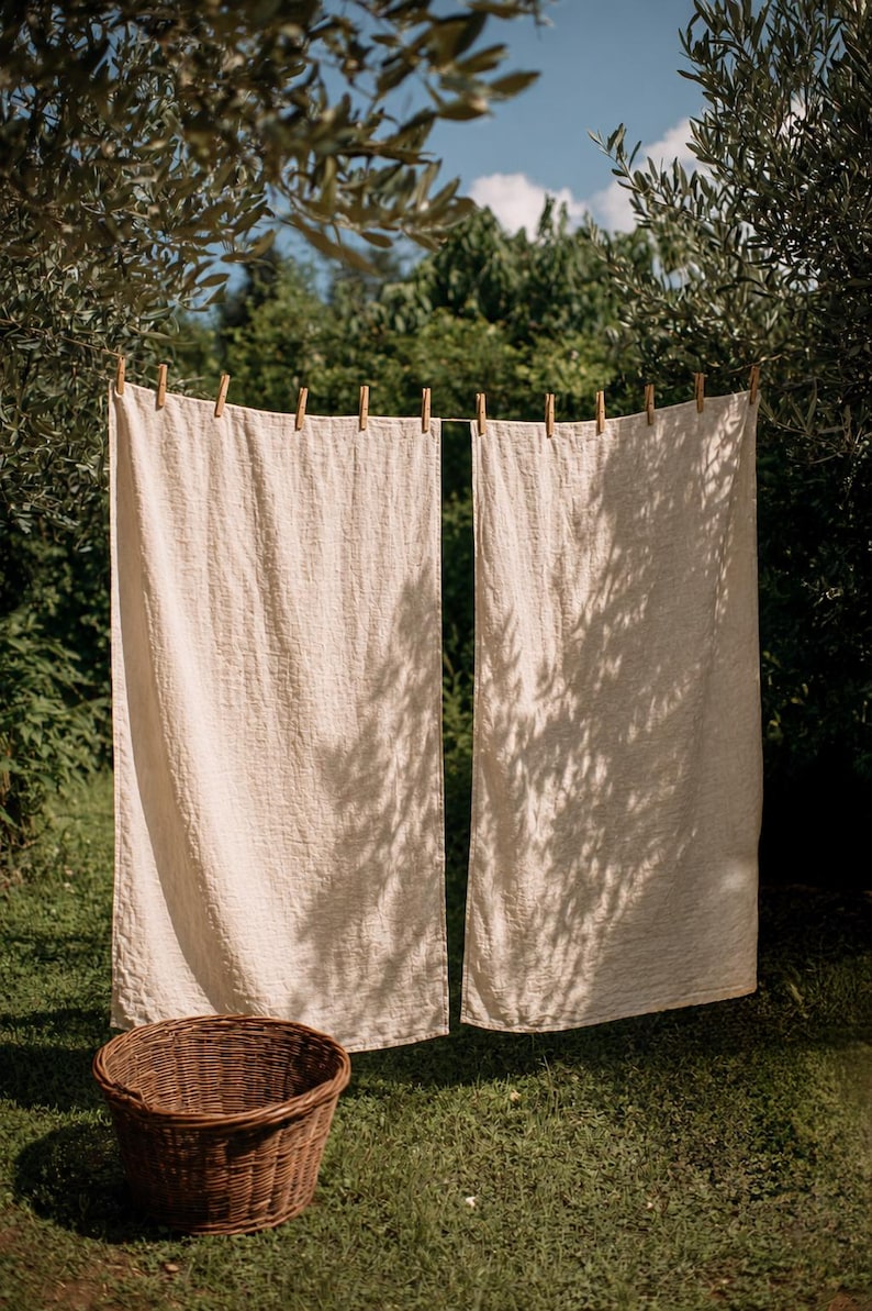 May include: Two rectangular, off-white linen towels hang side-by-side from a horizontal clothesline secured with wooden clothespins. A round, woven brown wicker basket sits on the ground below the hanging fabric, which features a visible weave.
