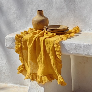 May include: A mustard-yellow linen table runner with a ruffled edge, stacked plates, and a textured, tan-coloured vase. The items are arranged on a white surface against a white wall. The plates and vase are a similar colour.