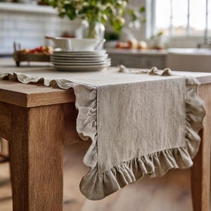 May include: A neutral-toned linen table runner with ruffled edges. The runner is draped over a wooden table, showcasing its textured fabric and decorative trim. The kitchen setting includes dishes and flowers.