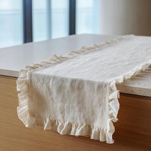 May include: A cream linen table runner with ruffled edges. The rectangular runner sits on a light-coloured countertop. The ruffles provide a decorative element to the simple design. The runner is made of a textured fabric.