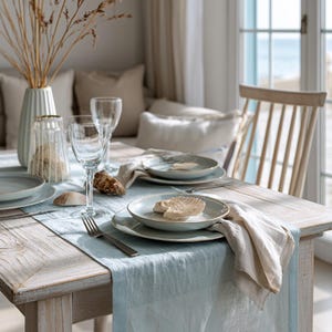 May include: A coastal-themed dining table setting with light blue linen runner, plates, and napkins. The table is set with wine glasses, a vase with dried grasses, and decorative seashells. The wooden chair and table have a weathered appearance.