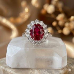 Puede incluir: Un anillo de plata con una gran gema de rubí ovalada rodeada por un halo de diamantes claros más pequeños. El anillo está colocado sobre una superficie geométrica blanca, con un fondo borroso.