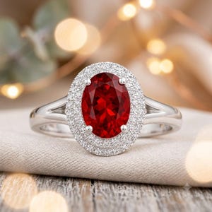 May include: A silver-toned ring featuring a large, oval-cut deep red gemstone at the centre. The stone is surrounded by a double halo of small, clear, sparkling stones. The band is made of a polished metallic material and splits into two sections.