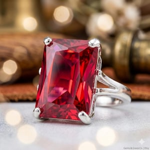 Op de afbeelding: Een zilveren ring met een grote, rechthoekige, gefacetteerde rode edelsteen. De edelsteen is gezet met vier zilveren pootjes. De ringband heeft een gespleten ontwerp. De achtergrond is wazig, met warme tinten en bokeh.