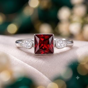 Puede incluir: Un anillo de tono plateado que presenta una gran piedra preciosa de color rojo intenso con corte cuadrado en el centro. La banda está acentuada a cada lado por una piedra transparente más pequeña en forma de pera, creando un diseño de tres piedras con acabado pulido.