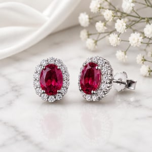 May include: A pair of stud earrings featuring oval-cut, deep red gemstones surrounded by a halo of small, clear stones. The earrings are set in a silver-colored metal and rest on a white surface, with white flowers in the background.