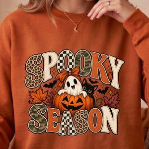 Op de afbeelding: Een oranje sweatshirt met lange mouwen en de tekst "SPOOKY SEASON" in grote, gedessineerde letters. De afbeelding bevat een wit spookje met een strik, een zwarte kat, een pompoen en herfstbladeren met kleine vleermuizen op de achtergrond.