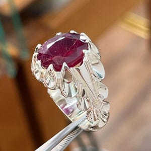 Peut inclure: Une bague en argent ornée d'une grande pierre précieuse rouge foncé à facettes, fixée par des griffes. L'anneau de la bague présente un motif répétitif en forme de feuilles ou de pétales qui s'étend de la base du sertissage le long de l'anneau.