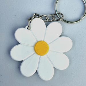 May include: A white daisy-shaped keyring with a yellow centre and silver chain and ring. The daisy has detailed petals and a three-dimensional design. A charming accessory.