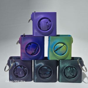 May include: A stack of six colorful, 3D-printed puzzle boxes. Each cube-shaped box has a circular opening revealing a complex, spherical puzzle. The boxes are in shades of purple, blue, and green, with keyrings attached.