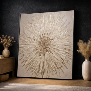 May include: Large abstract wall art with a black frame. The artwork features a central burst design in shades of gold and beige, creating a textured, dimensional effect. The piece is displayed in a room with neutral decor, including vases and a wooden cabinet.